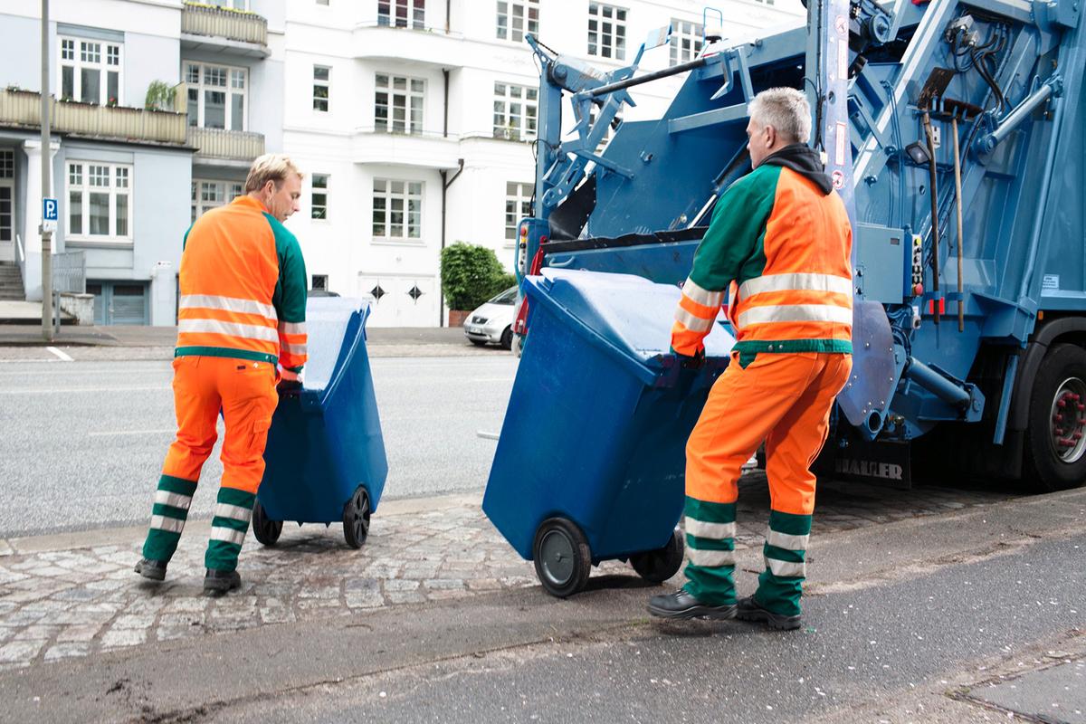Öffnet eine Lightbox: Zwei Müllmänner heben jeweils eine entleerte blaue Mülltonne eine Bordsteinkante hinauf. Ihr Müllwagen hält auf einer Parkfläche neben dem Bordstein.