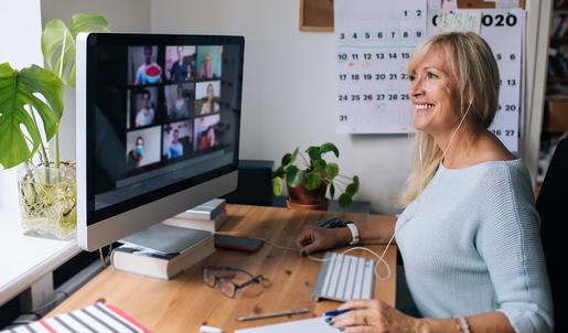 Eine Frau sitzt an einem Schreibtisch vor einem Computerbildschirm. Sie befindet sich gerade in einer Zoom-Konferenz und trägt daher Headphones. Um sie herum stehen Topfpflanzen, im Hintergrund hängt ein Kalender.