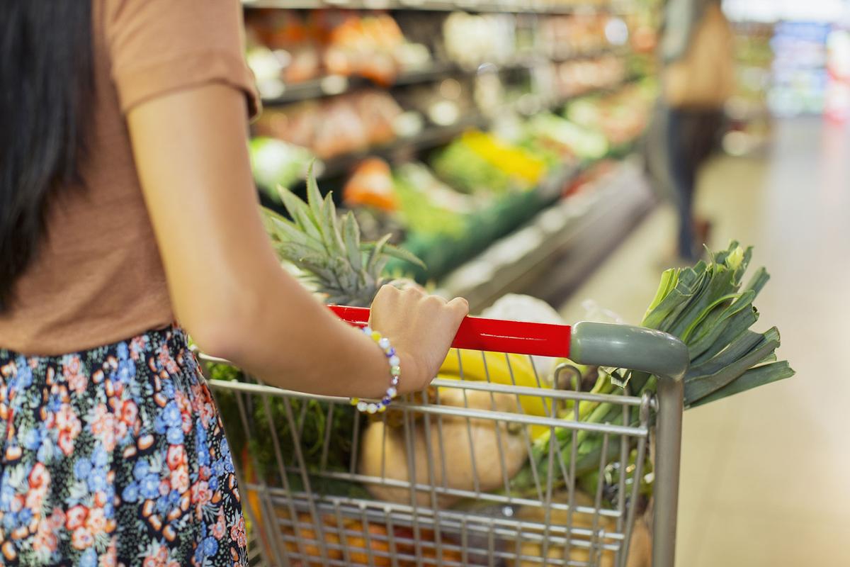 Großansicht: Eine Frau schiebt einen mit Lebensmitteln bestückten Einkaufswagen durch die Gemüseabteilung eines Supermarkts.
