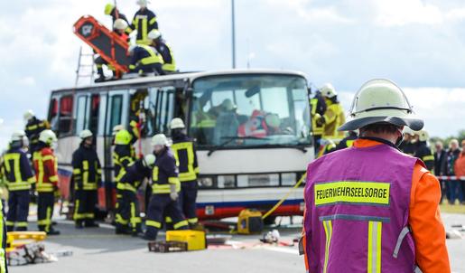 Im Hintergrund steht eine Gruppe von Feuerwehrleuten an einem weißen Bus. Im Vordergrund steht ein Feuerwehrmann mit einer lilafarbenen Weste mit der Aufschrift "Feuerwehrseelsorge". 