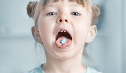 Ein Kind öffnet den Mund und streckt die Zunge raus. Auf seiner Zunge liegt ein blaues Medikament.