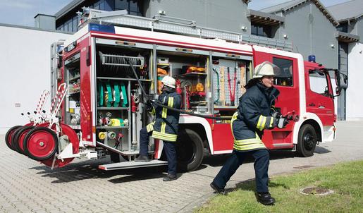 Zwei Feuerwehrleute in Schutzkliedung ziehen einen Schlauch aus einem geöffneten Feuerwehr-LKW