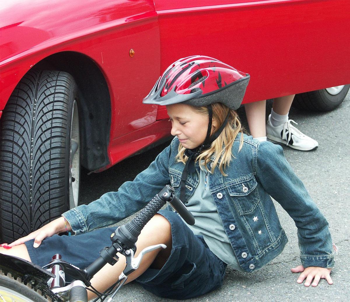 Mädchen mit Helm sitzt weinend auf der Straße. Vor ihr liegt ein Fahrrad, hinter ihr steigt jemand aus einem Auto. 