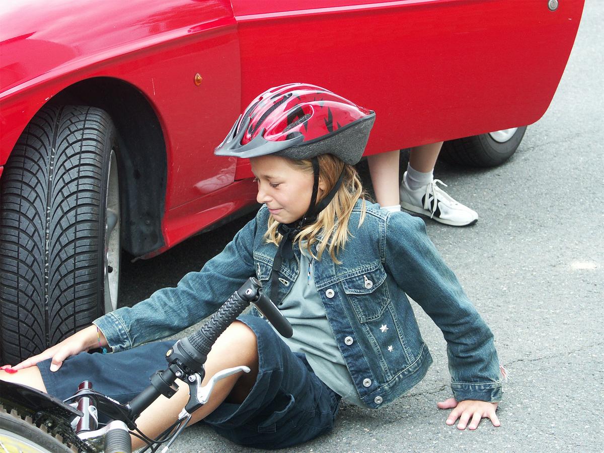 Mädchen mit Helm sitzt weinend auf der Straße. Vor ihr liegt ein Fahrrad, hinter ihr steigt jemand aus einem Auto. 