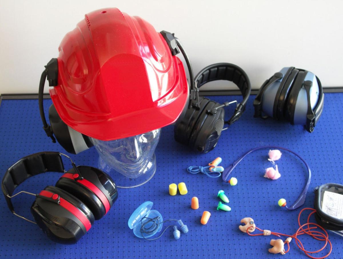 Großansicht: Auf einem Tisch liegen verschiedene Sorten an Gehörschutz: Otoplastik, Ohrstöpsel, Kapselgehörschutz und Helme mit integriertem Gehörschutz.