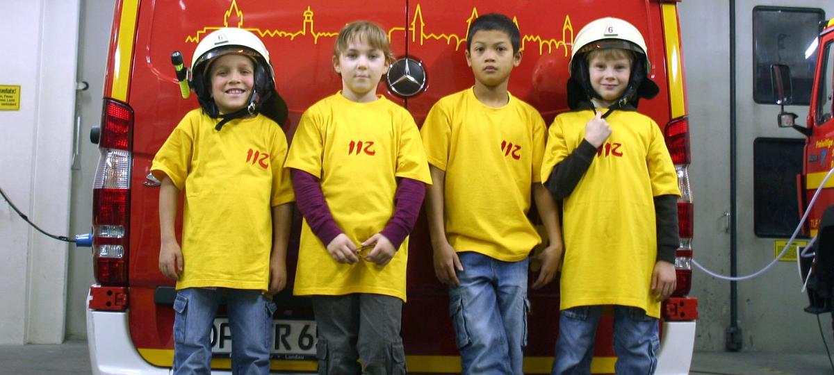Vier Kinder lehnen an der Rückseite eines Feuerwehrwagens. Zwei von ihnen tragen Feuerwehrhelme.