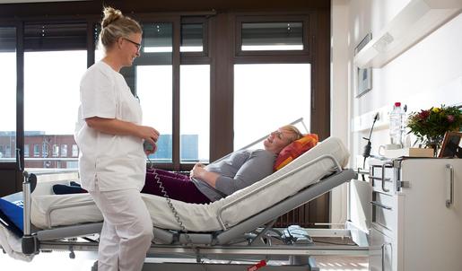 Krankenschwester stellt das Rückenteil eines Krankenhausbettes höher, eine Patientin liegt darin.