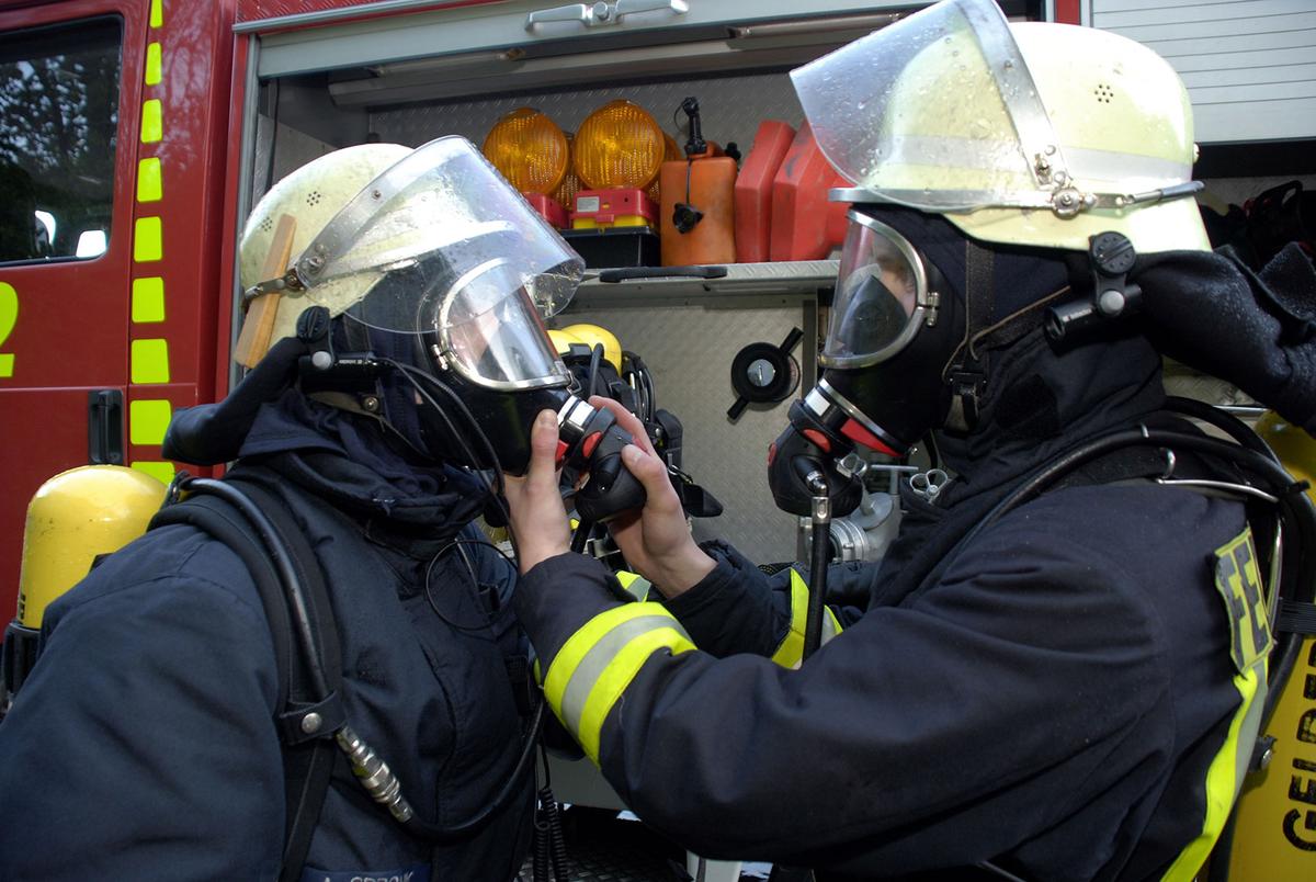 Öffnet eine Lightbox: Zwei Feuerwehrmänner mit aufgesetzten Helmen und Atemschutzgeräten stehen vor einem Feuerwehrfahrzeug. Der eine überprüft die Maske des anderen.