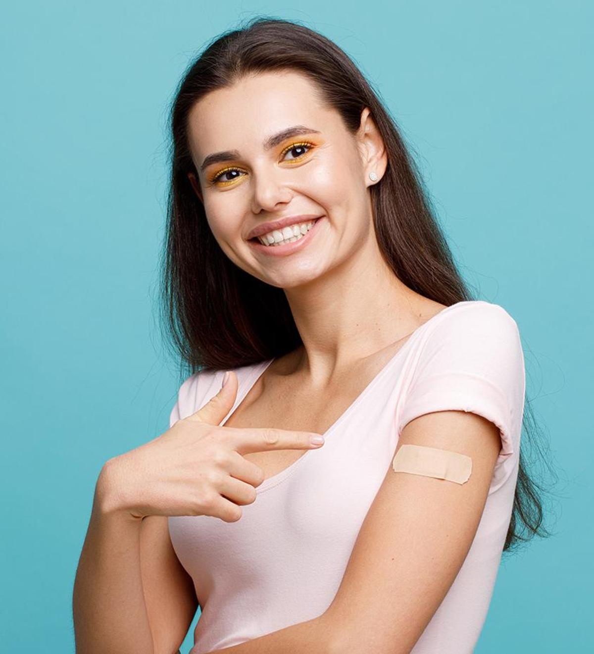 Großansicht: Eine junge Frau verweist lächelnd auf das Impfpflaster an ihrem linken Arm.