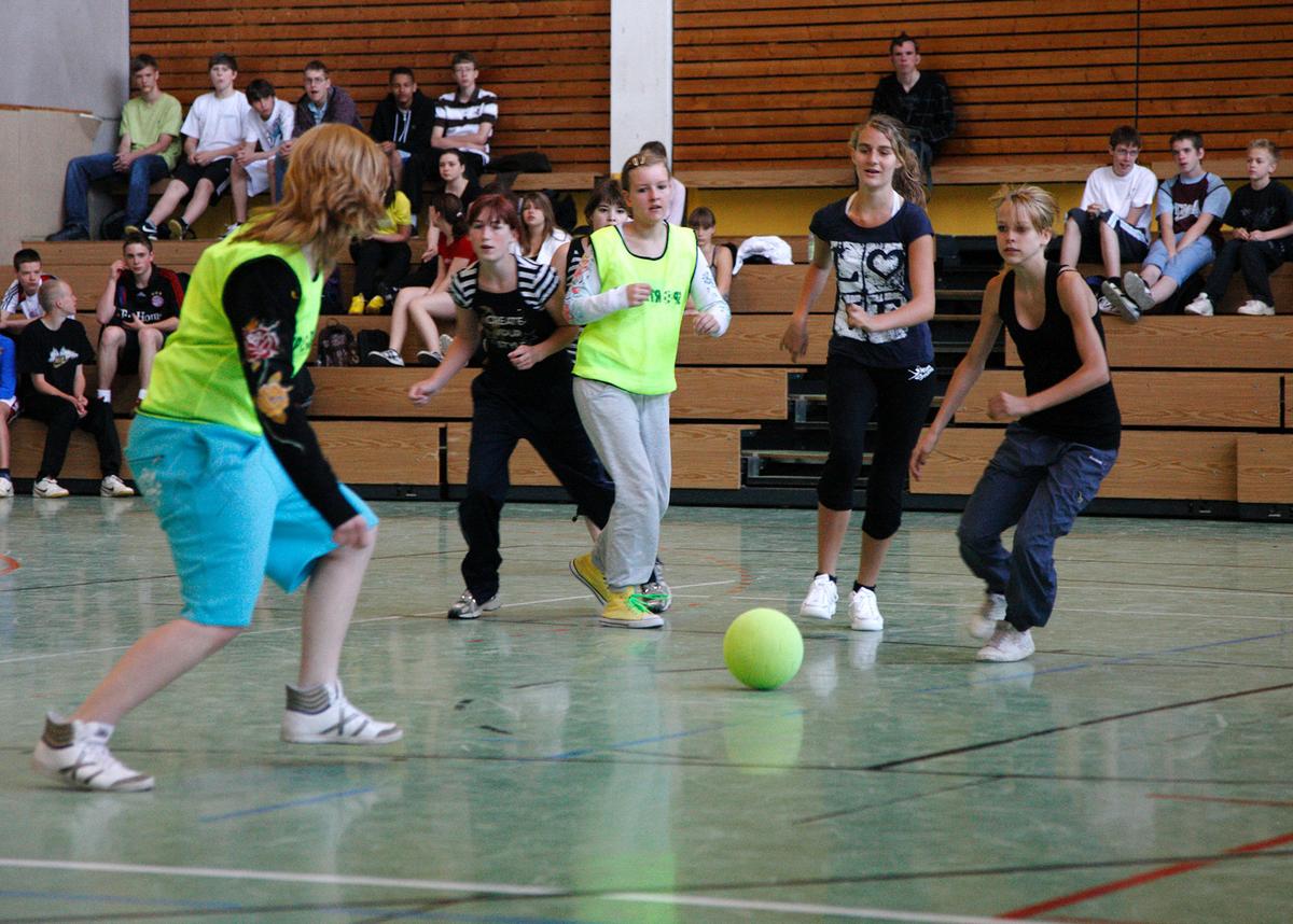 Großansicht: Schülerinnen spielen in einer Halle Fußball. Eins der Teams trägt gelbe Leibchen. Schüler schauen ihnen von den Tribünen aus zu.