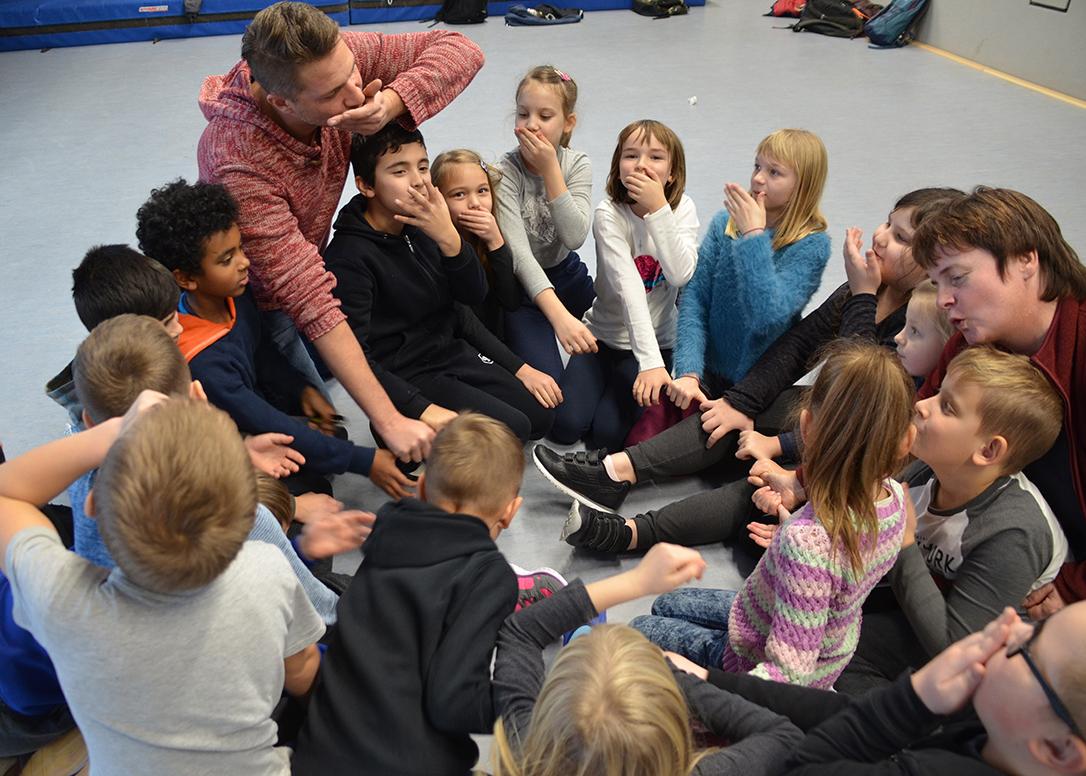 Der Projektleiter und eine Gruppe Grundschulkinder sitzen in einer Sporthalle eng beieinander in einem Kreis. Sie halten die rechte Hand in die Mitte des Kreises, und die linke vor den Mund.