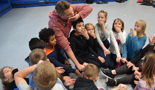 Der Projektleiter und eine Gruppe Grundschulkinder sitzen in einer Sporthalle eng beieinander in einem Kreis. Sie halten die rechte Hand in die Mitte des Kreises, und die linke vor den Mund.