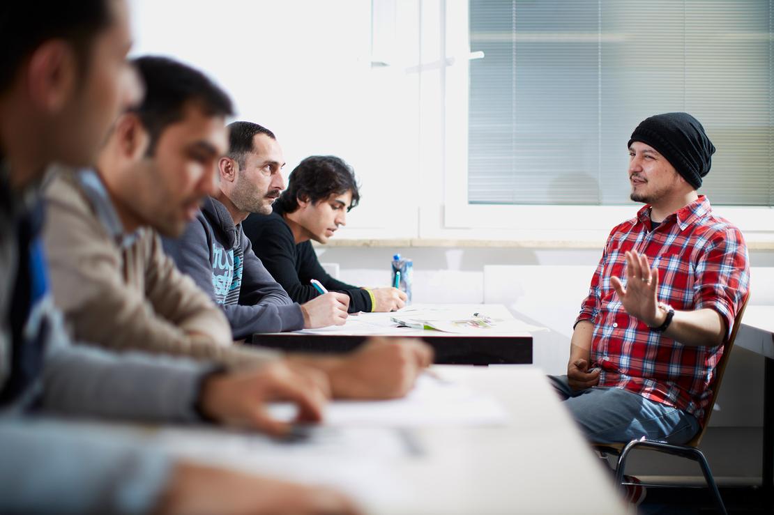 Vier m&auml;nnliche Gefl&uuml;chtete sitzen w&auml;hrend einer Schulungssituation an Schreibtischen und schauen zu einem gestikulierenden Lehrer