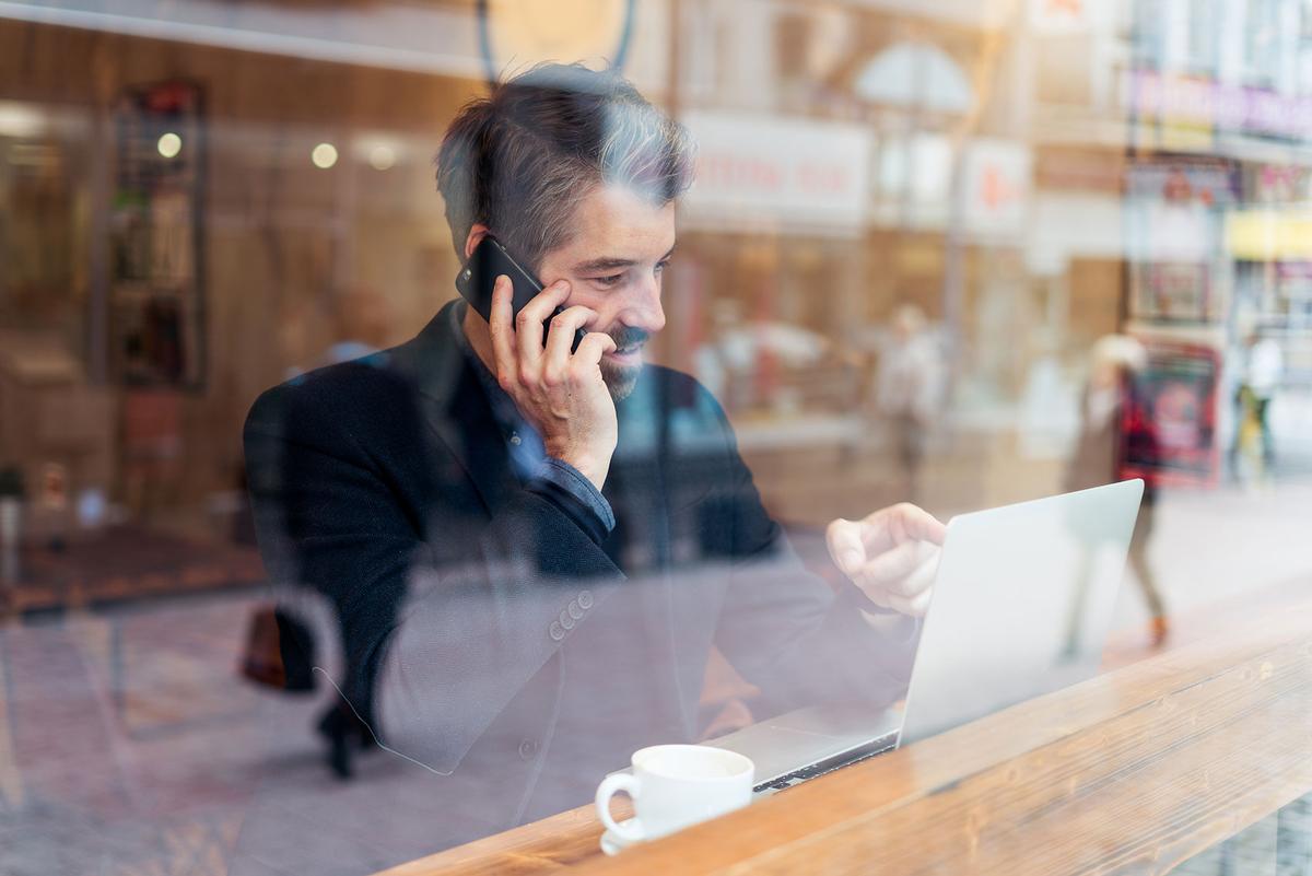 Ein Mann schaut in einem Café auf ein Tablet, während er mit dem Handy telefoniert. Neben ihm steht eine Tasse Kaffee.