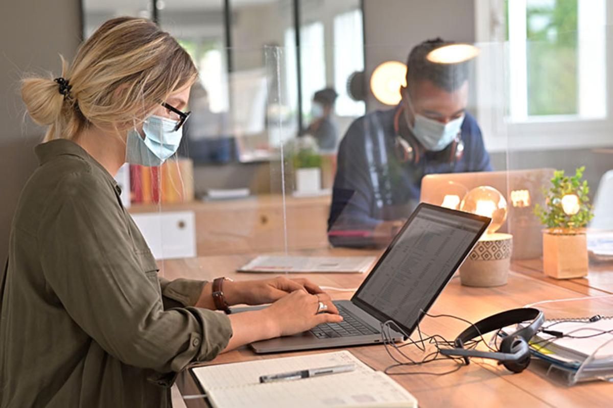 Öffnet eine Lightbox: Personal bei der Arbeit im Gemeinschaftsbüro. Im Vordergrund sitzen zwei Personen an ihren Schreibtischen und arbeiten mit medizinischer Maske und Trennschtuz an ihren Laptops.
