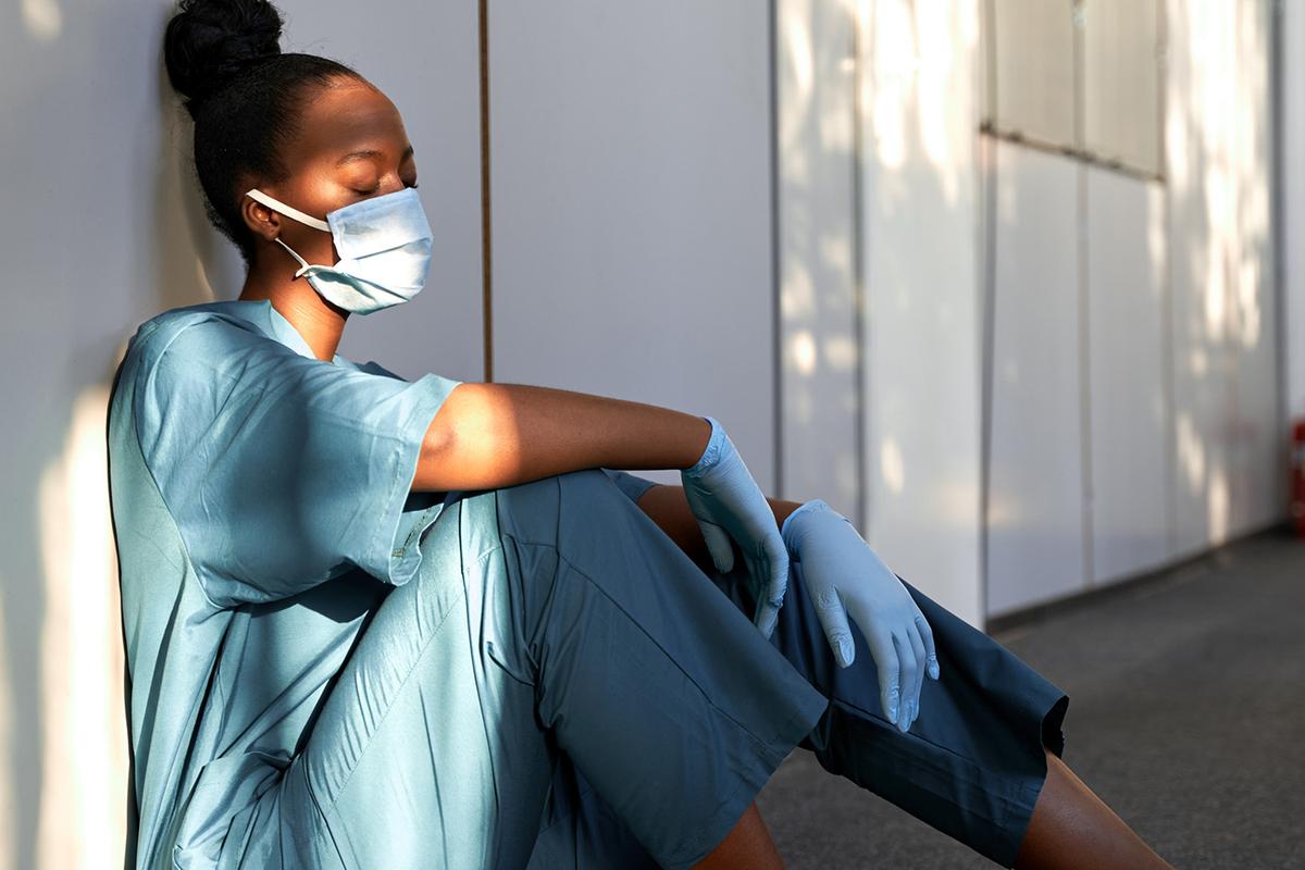 Erschöpfte Krankenschwester mit Maske und Einmalhandschuhen sitzt angelehnt an eine Wand in einem Flur.