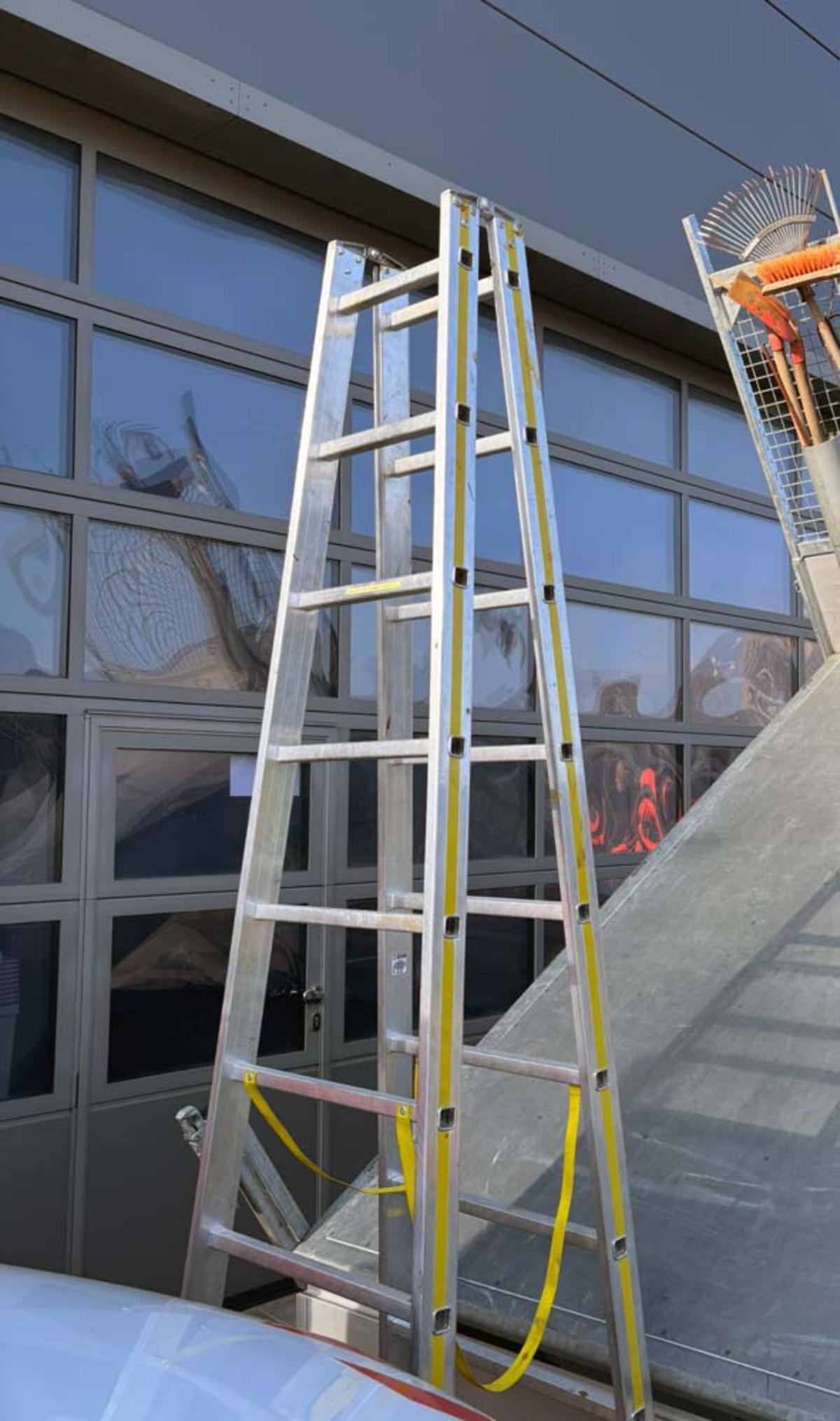&Ouml;ffnet eine Lightbox: Eine aufgestellte Aluminiumleiter ohne Trittstufen (Multifunktionsleiter) mit gelben Sicherheitsb&auml;ndern steht vor der gro&szlig;en Glasfront und dem Sektionaltor eines modernen, grauen Industriegeb&auml;udes. Im Vordergrund sind Teile eines wei&szlig;en Fahrzeugs und eines grauen Objekts zu sehen.