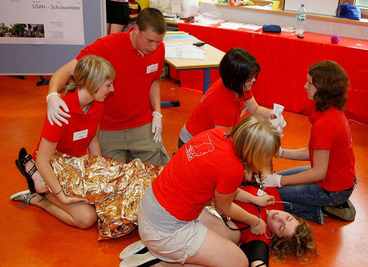 Öffnet eine Lightbox: Mitglieder eines Schulsanitätsdienst nehmen an einer Ersten Hilfe Übung teil. Eine Teilnehmerin wurde mit einer Rettungsdecke zugedeckt und ihre Beine nach oben gelegt. 