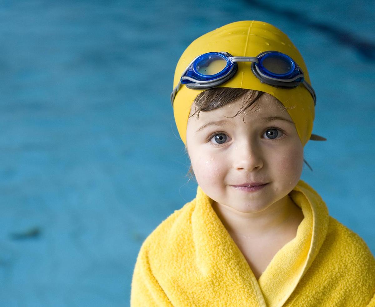 Kind mit Badekappe, Schwimmbrille und Handtuch steht vor einem Schwimmbecken.