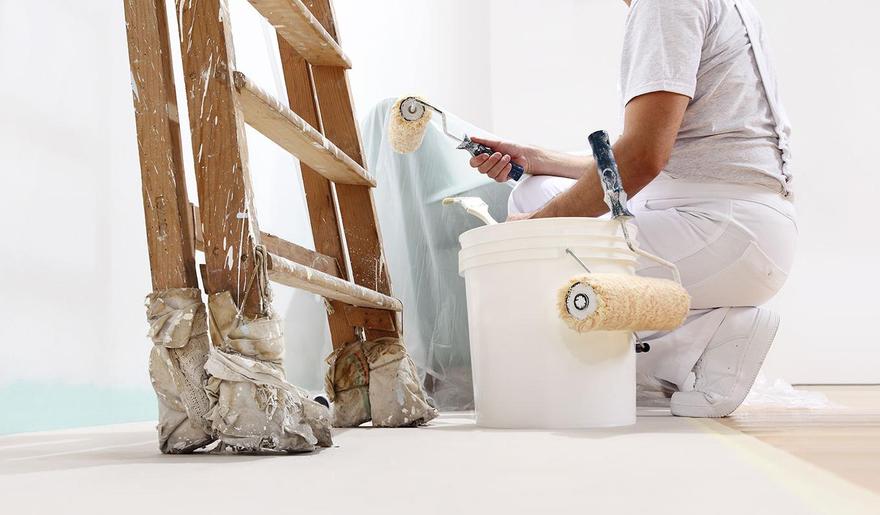Ein hell gekleideter Mann ist damit beschäftigt, eine Wand mit einem Rollpinsel weiß zu streichen. Um ihn herum stehen eine mit Farbspritzern befleckte Leiter, ein Farbeimer sowie ein mit Plastikfolie abgedecktes Möbelstück.