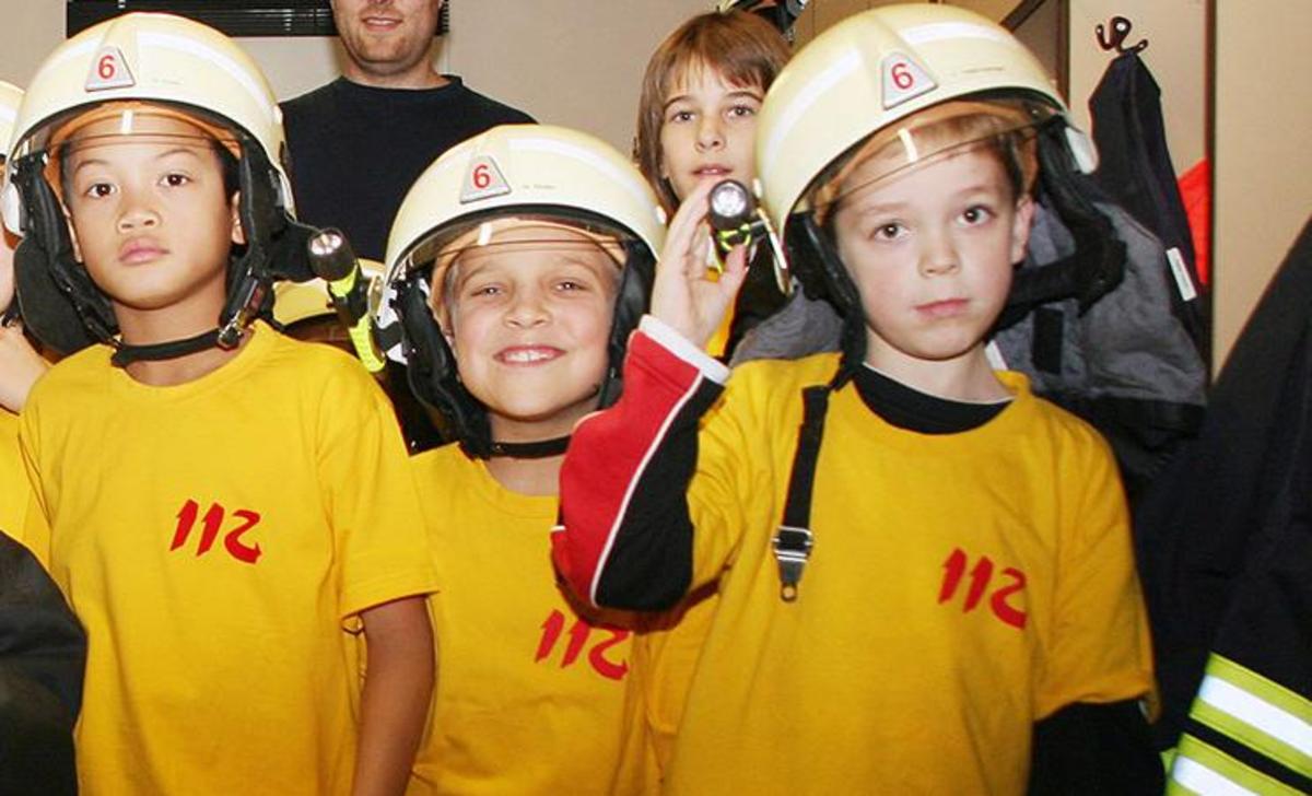 Eine Kindergruppe der Freiwilligen Feuerwehr steht im Ankleideraum einer Feuerwache. Einige tragen Brandbekämpfungs-Überjacken, andere Feuerwehrhelme.