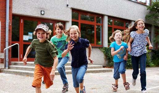 Fünf Schulkinder stürmen vom Schulgebäude weg über einen Schulhof. Sie wirken fröhlich.