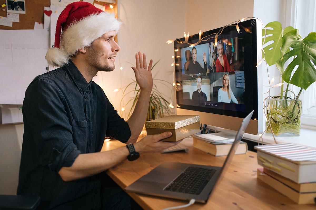 Großansicht: Virtuelle Meeting-Konferenz von zu Hause. Junger Mann, der eine rote Weihnachtsmannmütze trägt und eine Geschenkbox hält.