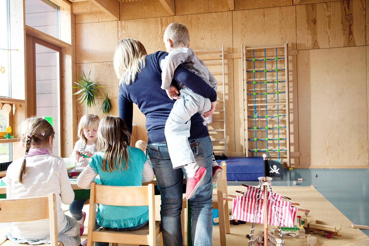 Großansicht:  In einem Bewegungsraum im Kindergarten trägt eine Erzieherin ein Kind auf der Hüfte, dabei beugt sie sich zu Kindern herunter, die an Tischen sitzen und malen.