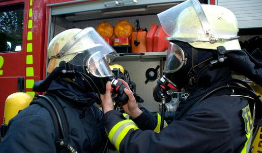 Zwei Feuerwehrm&auml;nner unter Atemschutz. Im Hintergrund befindet sich ein Feuerwehrfahrzeug.