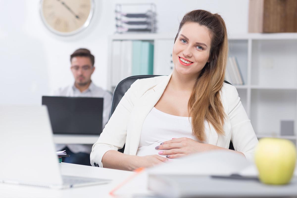 Öffnet eine Lightbox: Eine schwangere Frau sitzt im Büro an einem Laptop und lächelt in die Kamera.