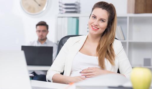 Eine schwangere Frau sitzt im Büro an einem Laptop und lächelt in die Kamera.