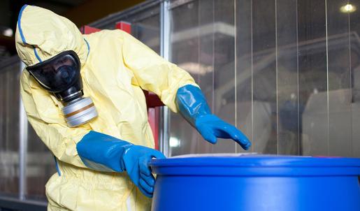 Eine Person in einem gelben Schutzanzug trägt blaue lange Gummihandschuhen und eine Atemschutzmaske, während sie an einem blauen Behältnis für Gefahrstoffe arbeitet.