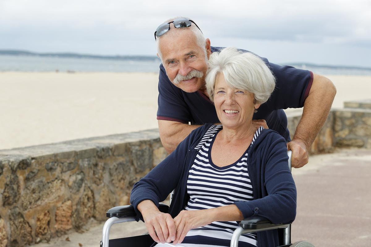 Großansicht: Ein älterer Herr mit Sonnenbrille schiebt eine in einem Rollstuhl sitzende ältere Dame einen gepflasterten Gehweg nahe eines Strands entlang. Beide wirken erheitert und lächeln in die Kamera.