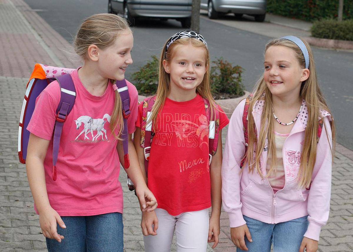 Öffnet eine Lightbox: Drei Schulmädchen gehen tagsüber einen Bürgersteig entlang. Alle drei tragen Schulranzen auf dem Rücken.