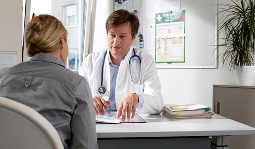 Ein Arzt sitzt in seiner Praxis an einem Schreibtisch und erläutert einer Patientin etwas.