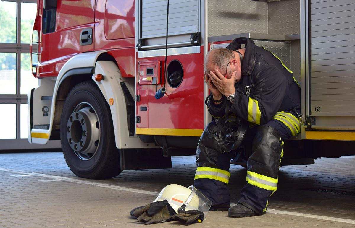 Öffnet eine Lightbox: Ein älterer Feuerwehrmann sitzt in Schutzkleidung auf der Trittstufe eines Feuerwehr-LKW und hält sich erschöpft beide Hände vor das Gesicht