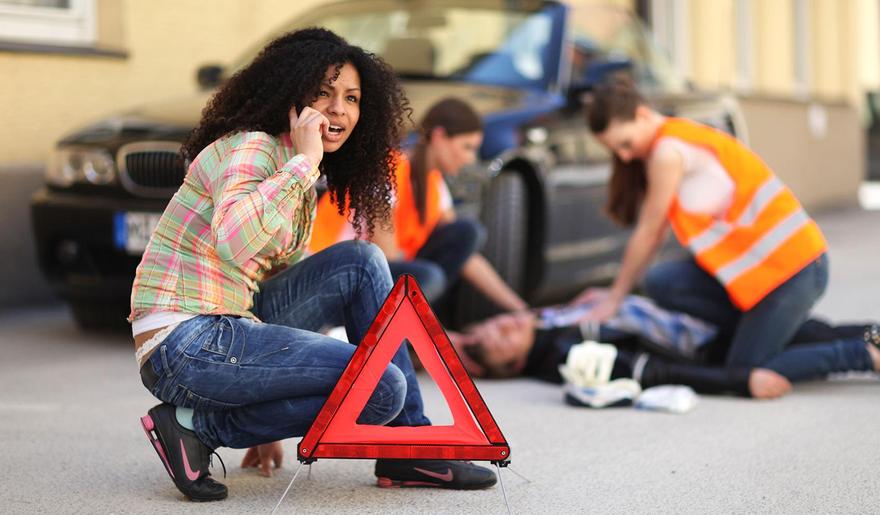 Eine Frau hockt an einer mit Warndreieck gesicherten Unfallstelle und setzt einen Notruf ab. Im Hintergrund wird vor einem Auto von zwei Frauen Erste Hilfe geleistet.