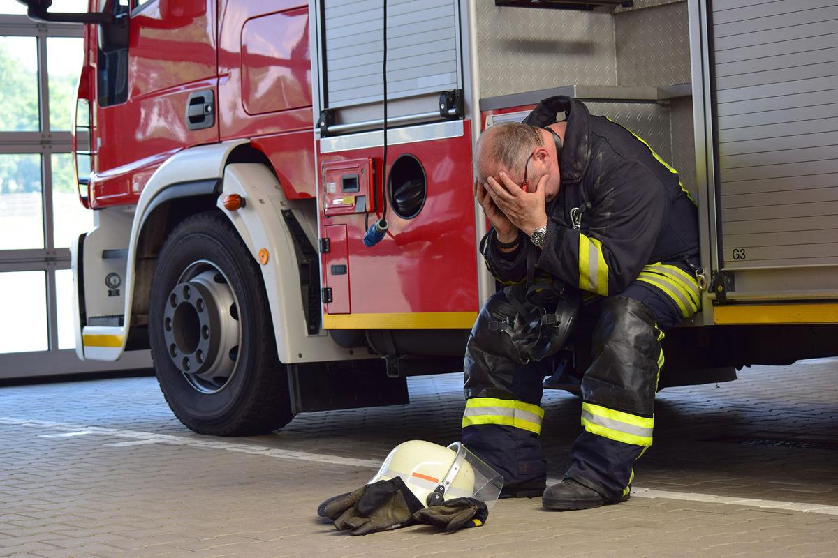 Ein Feuerwehrmann sitzt angelehnt an einem Einsatzfahrzeug und vergräbt sein Gesicht in den Händen. Vor ihm liegen Helm und Handschuhe.