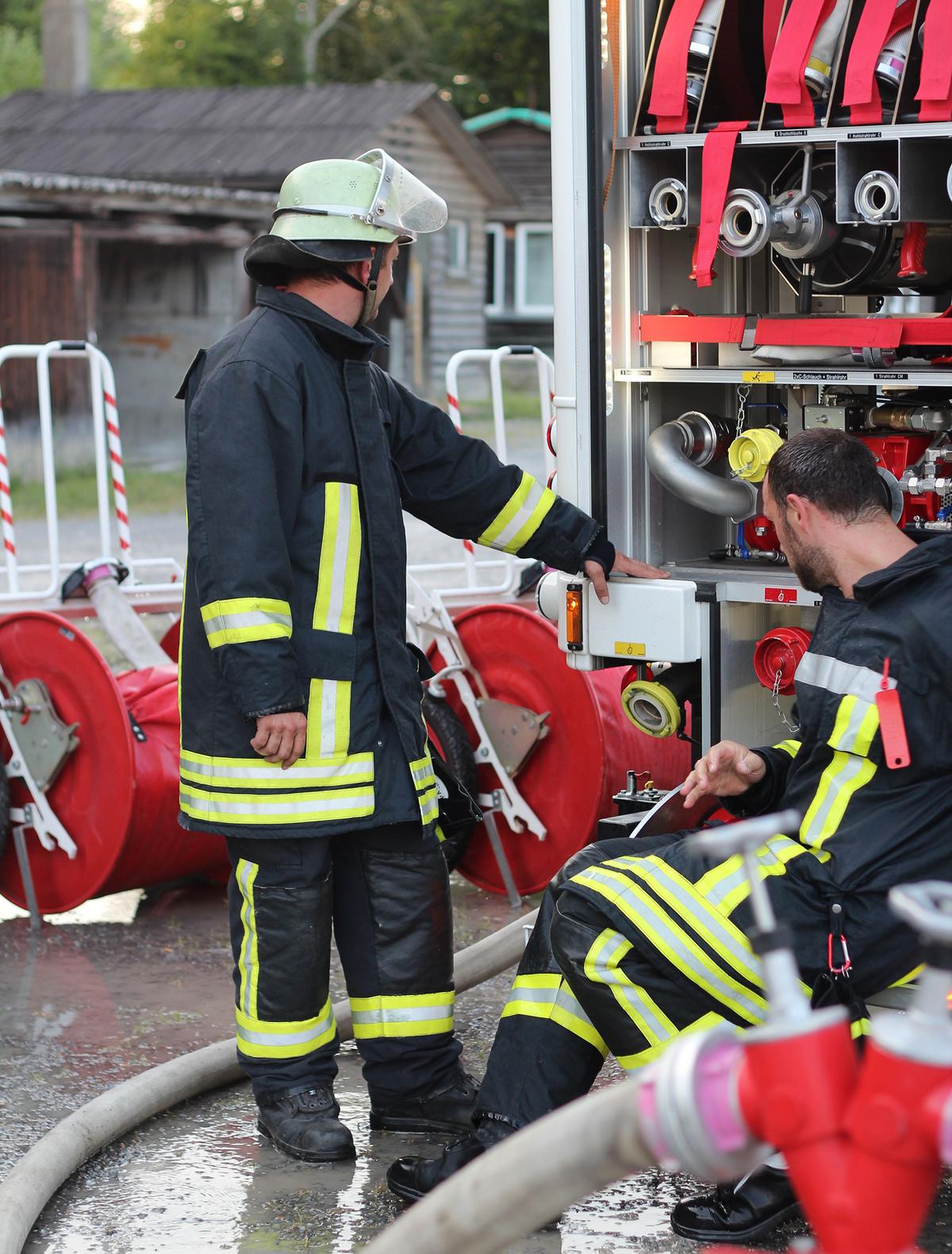 &Ouml;ffnet eine Lightbox: Zwei Feuerwehrm&auml;nner arbeiten an einem ge&ouml;ffneten Feuerwehrfahrzeug. Einer steht links und ber&uuml;hrt ein technisches Detail, der andere hockt rechts daneben. Im Hintergrund sind Schl&auml;uche und Geb&auml;udeteile zu sehen.
