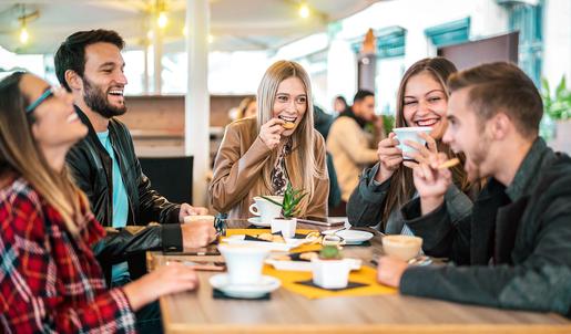 F&uuml;nf junge Personen sitzen um einen Tisch und trinken fr&ouml;hlich Kaffee.