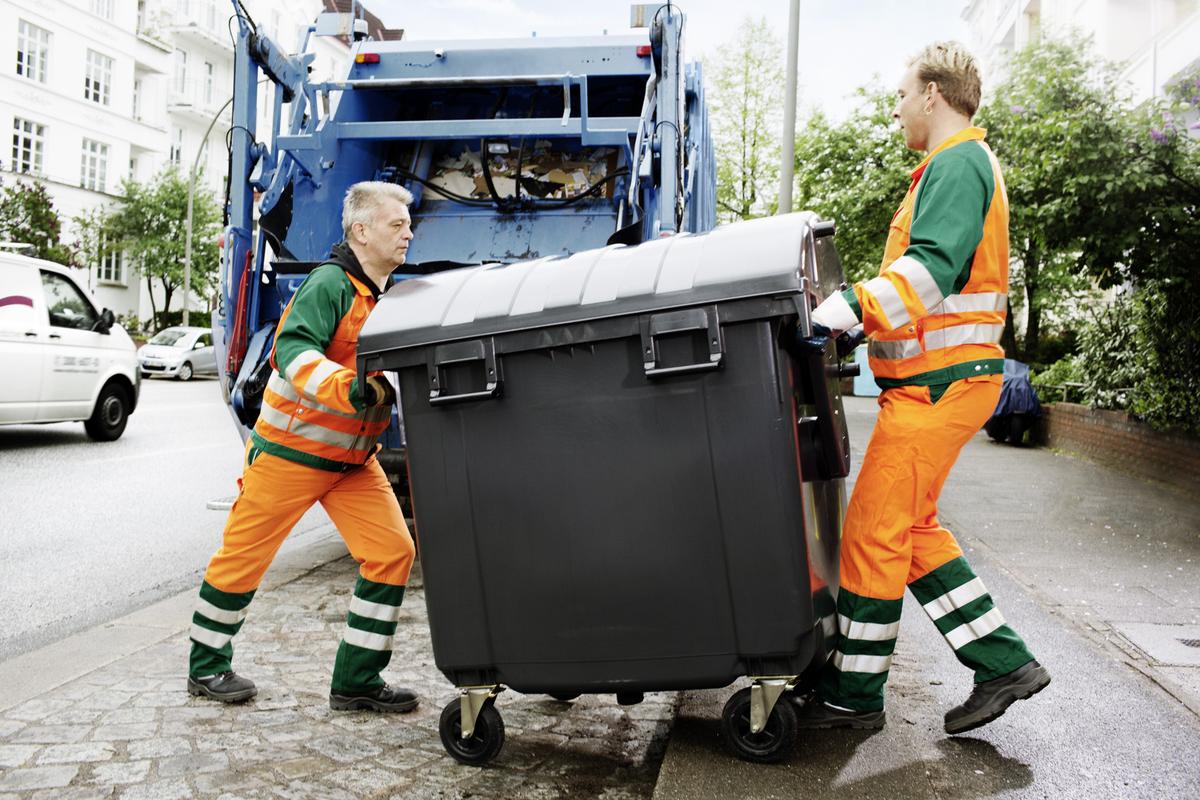 Öffnet eine Lightbox: Zwei Müllwerker schieben hinter einem Müllwagen eine grosse Mülltonne zurück auf einen Bürgersteig