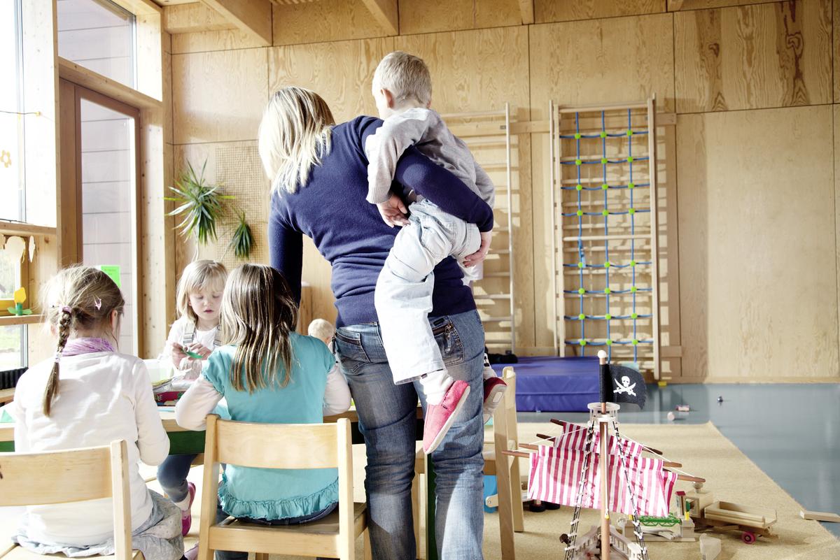 Eine Erzieherin mit Kind auf dem Arm steht an einem Tisch, an dem Kinder sitzen. 