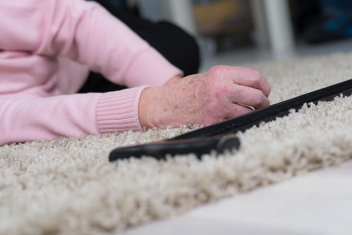 Eine alte Frau ist gestürzt und liegt auf einem Teppich. Sie hat die Hände vom Körper gestreckt und ein Gehstock liegt neben ihr. 