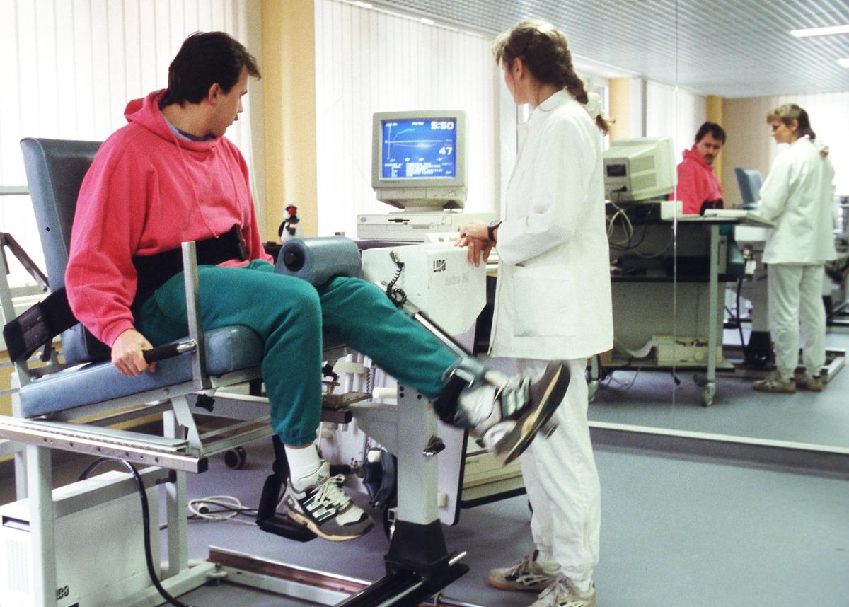 Großansicht: Ein Mann sitzt auf einem medizinischen Gerät. Eine medizinisch Angestellte steht daneben und überwacht die Übungen, die der Mann auf dem Gerät macht.