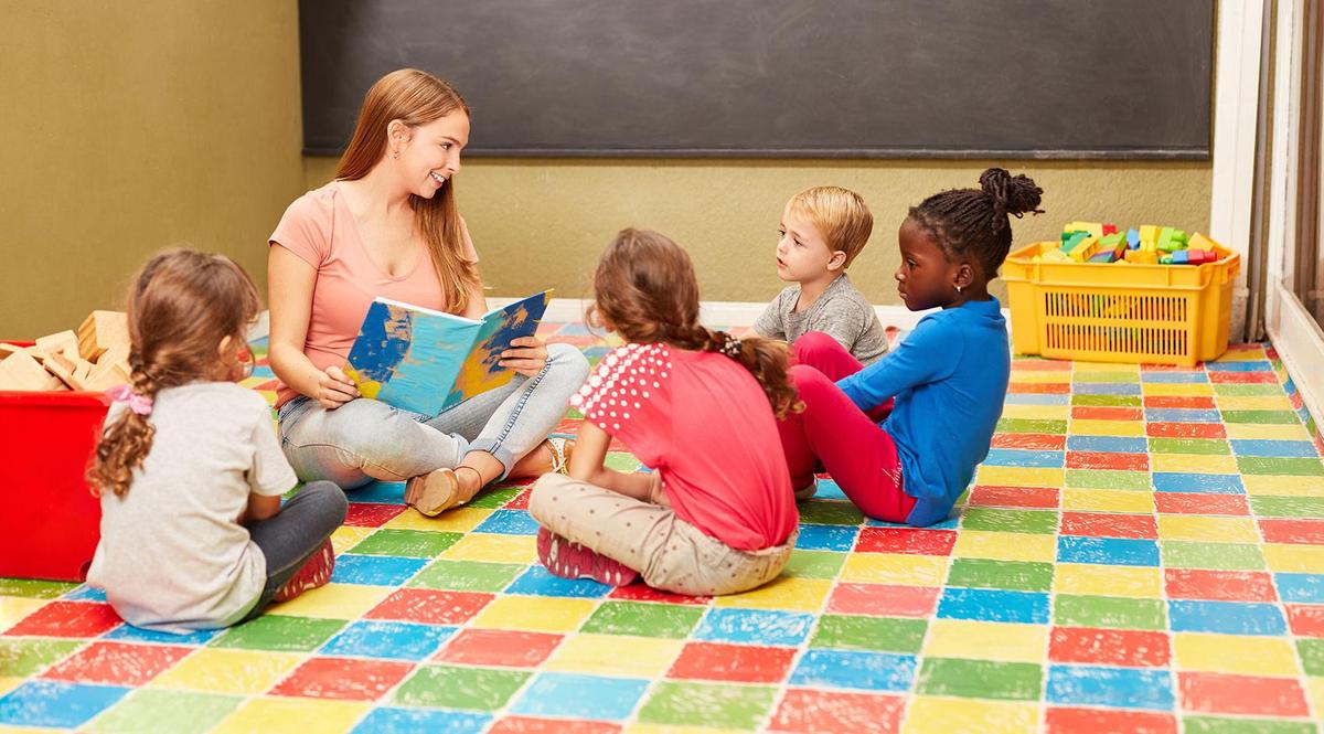 Eine Frau sitzt mit einer Gruppe von Kindern auf dem Boden und liest ihnen aus einem Buch vor.