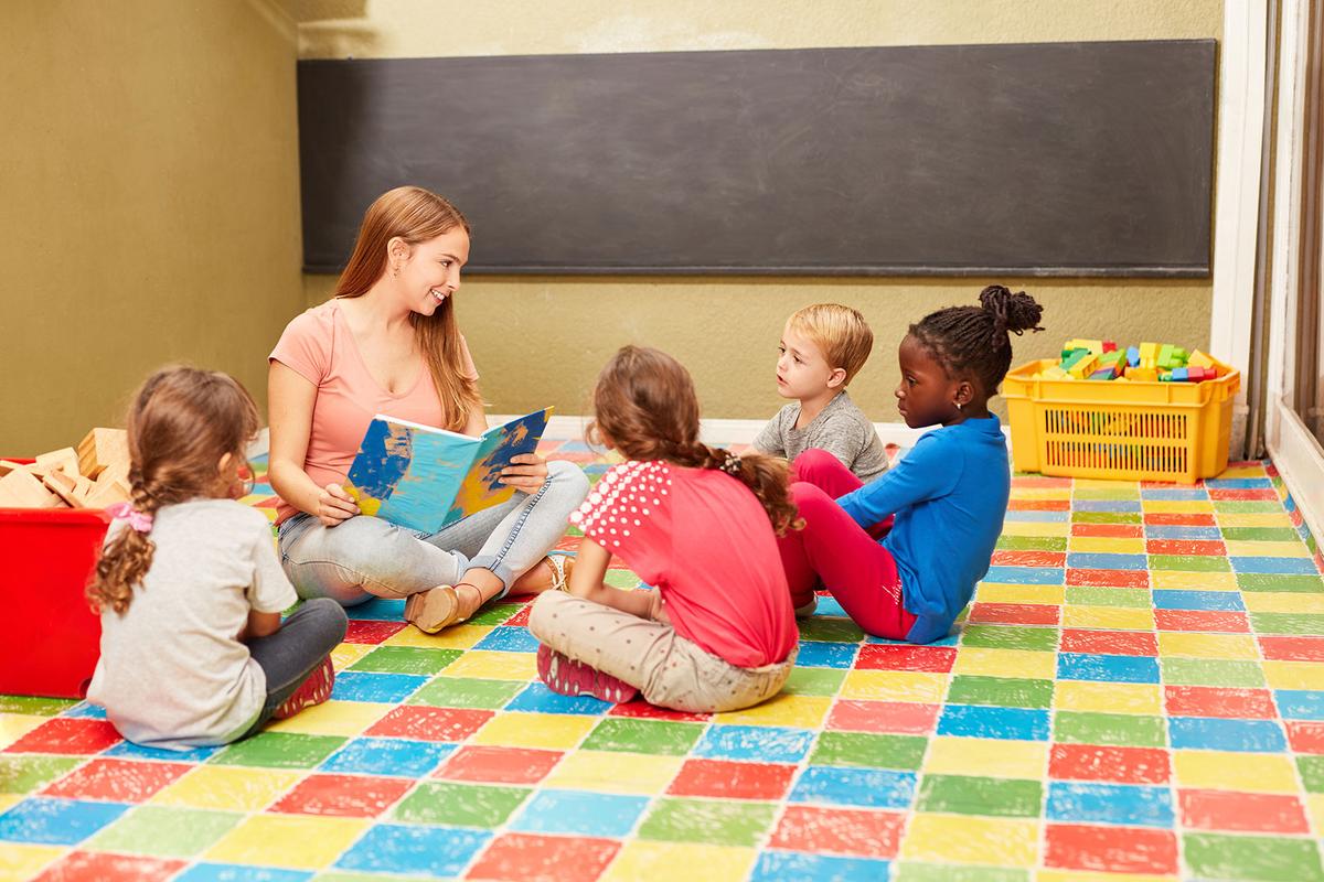Großansicht: Eine Frau sitzt mit einer Gruppe von Kindern auf dem Boden und liest ihnen aus einem Buch vor.