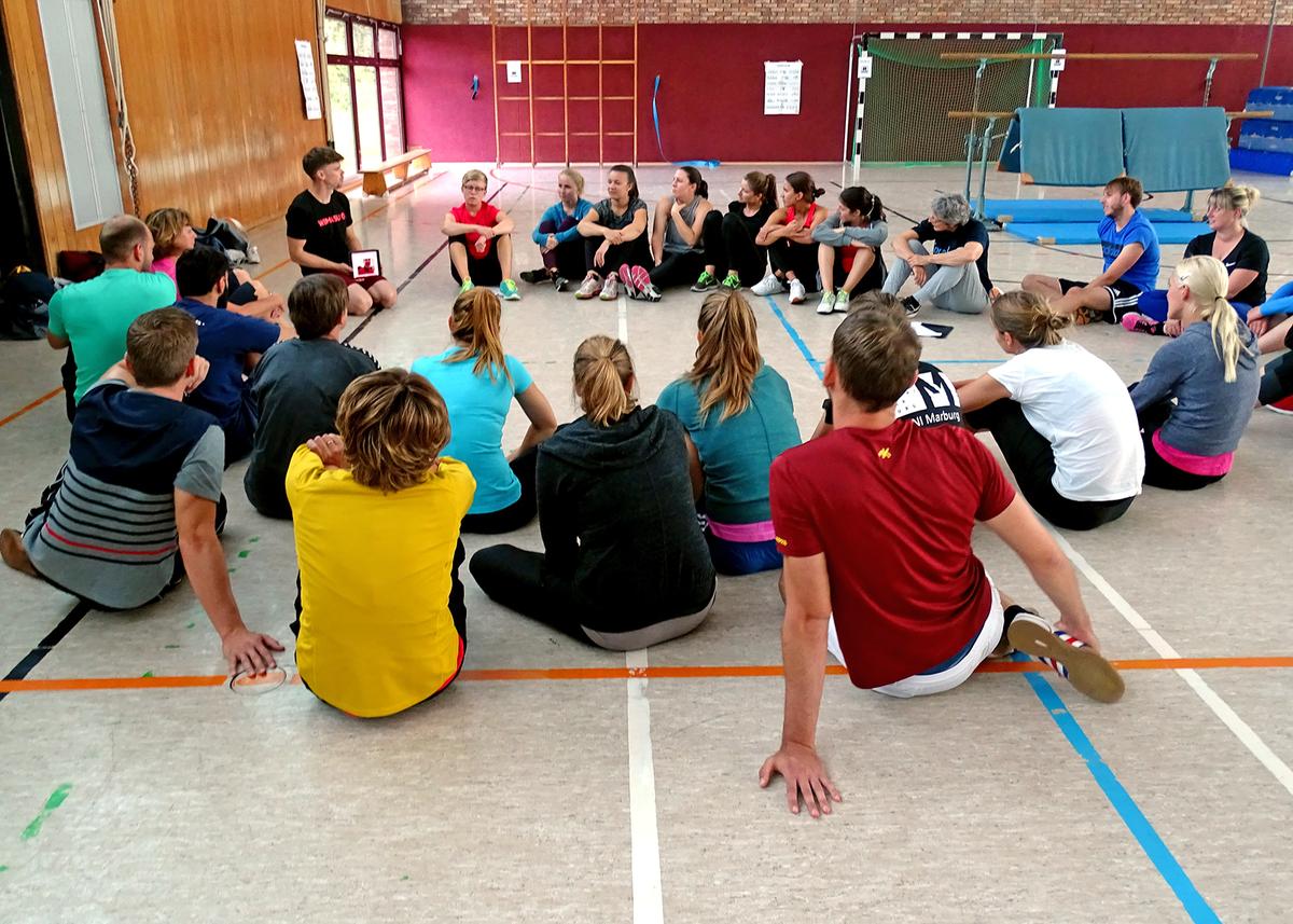 Öffnet eine Lightbox: Eine Gruppe Jugendlicher sitzt in einer Sporthalle im Kreis. Sie haben ihre Augen auf den Sportlehrer gerichtet, der ihnen etwas zu erklären scheint.