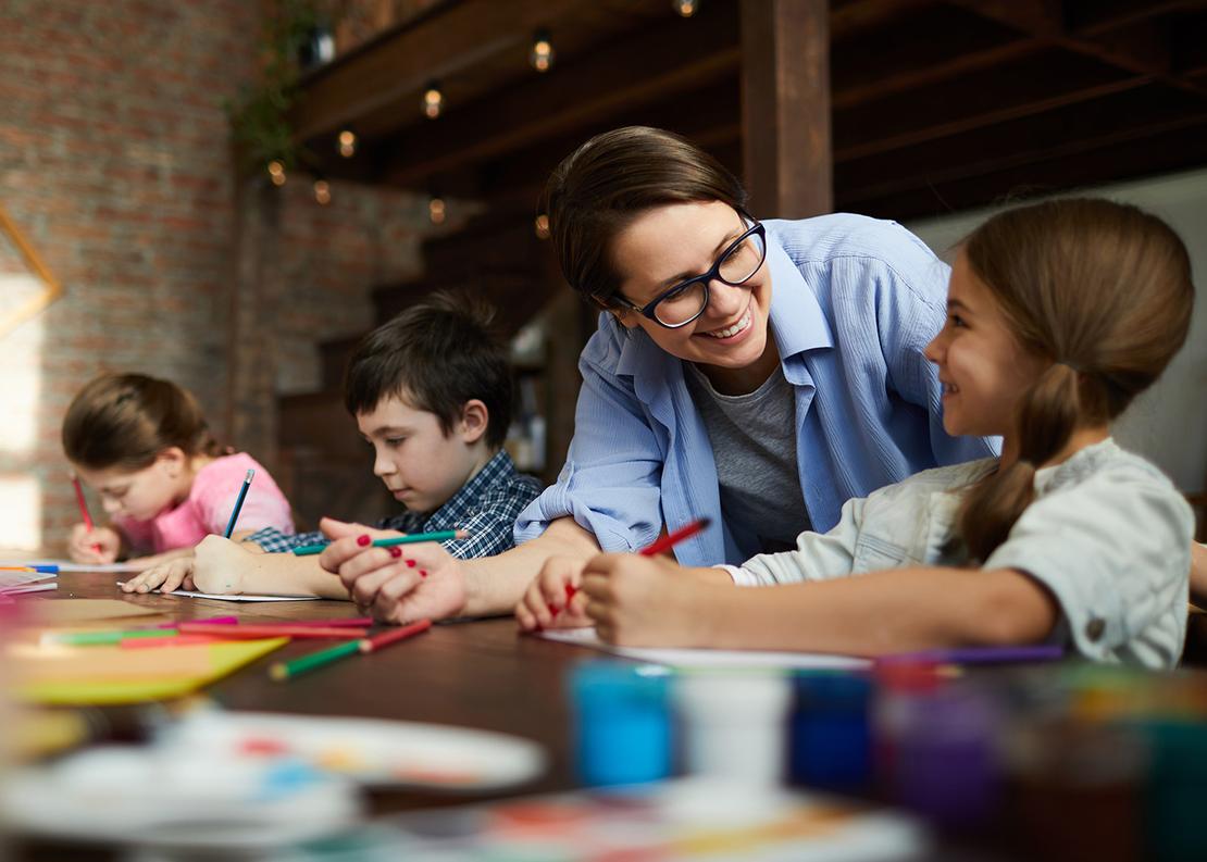 Drei Kinder zeichnen am Tisch in einer Kita, eine Betreuerin beugt sich lachend &uuml;ber sie