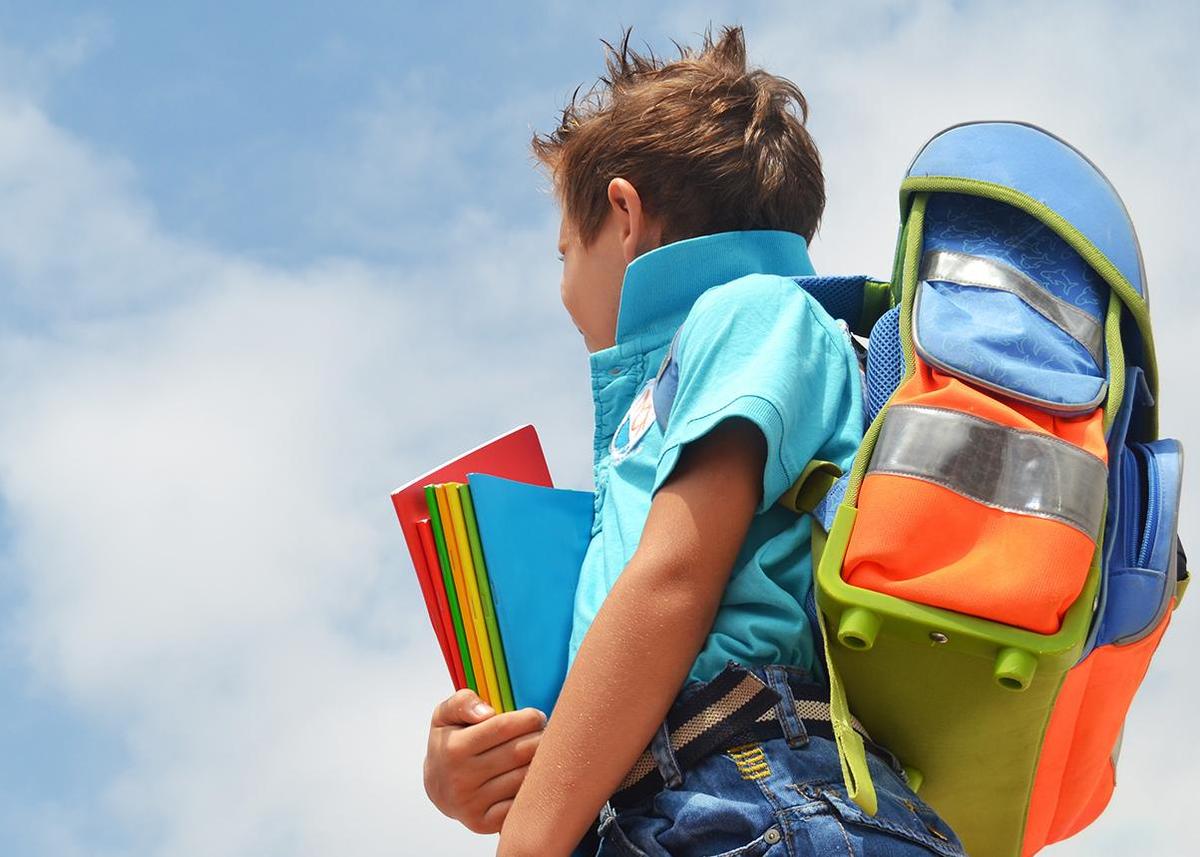 Ein Grundschulkind mit einem Schulranzen auf dem Rücken und einem Stapel Hefte in der Hand schaut nach oben in den blauen Himmel.
