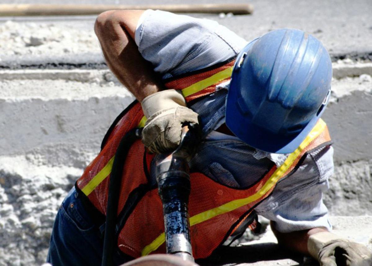 Großansicht: Ein Mann mit Schutzhelm, Warnweste und Schutzhandschuhen arbeitet auf einer Baustelle. 